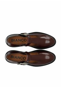 Rudos patento odos batai su uždara nosine, išpjovų dizainu, metaliniu sagties akcentu ir tekstūruotu padu. Logotipas matomas ant vidpadžio.