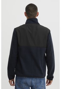 Man met kort haar en een litteken aan de achterkant van zijn hoofd, gekleed in een marineblauwe fleecejas met een zwart schouderstuk en blauwe spijkerbroek, van achteren gezien.