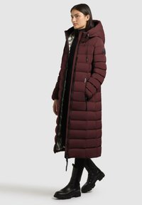 Bordeauxfarbene Pufferjacke mit Kapuze, langer Schnitt, gestepptes Design, mit silbernem Innenfutter und schwarzem Reißverschluss, getragen mit schwarzen kniehohen Stiefeln.