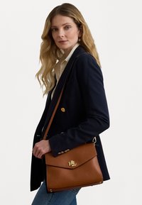 Brun läderhandväska med framficka och guldfärgade detaljer. Har en enkel design, axelrem och ett distinkt logotyp.