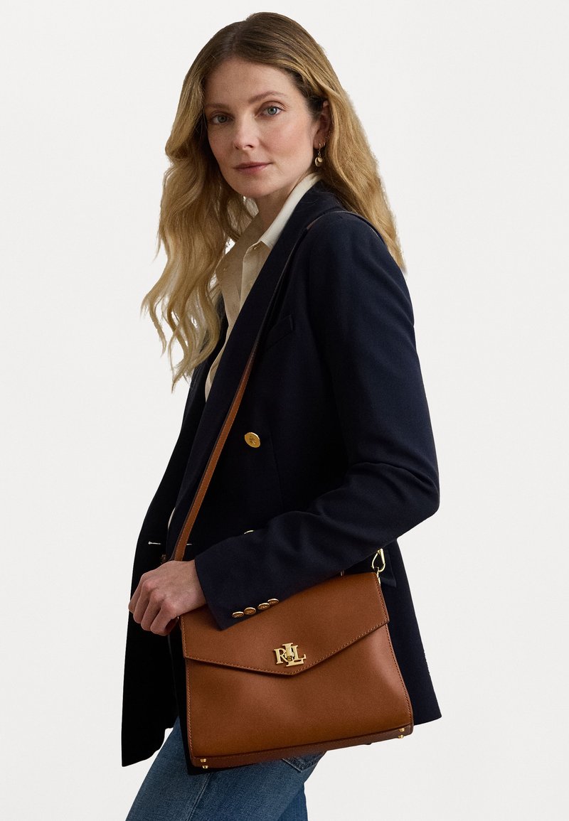 Brun läderhandväska med framficka och guldfärgade detaljer. Har en enkel design, axelrem och ett distinkt logotyp.