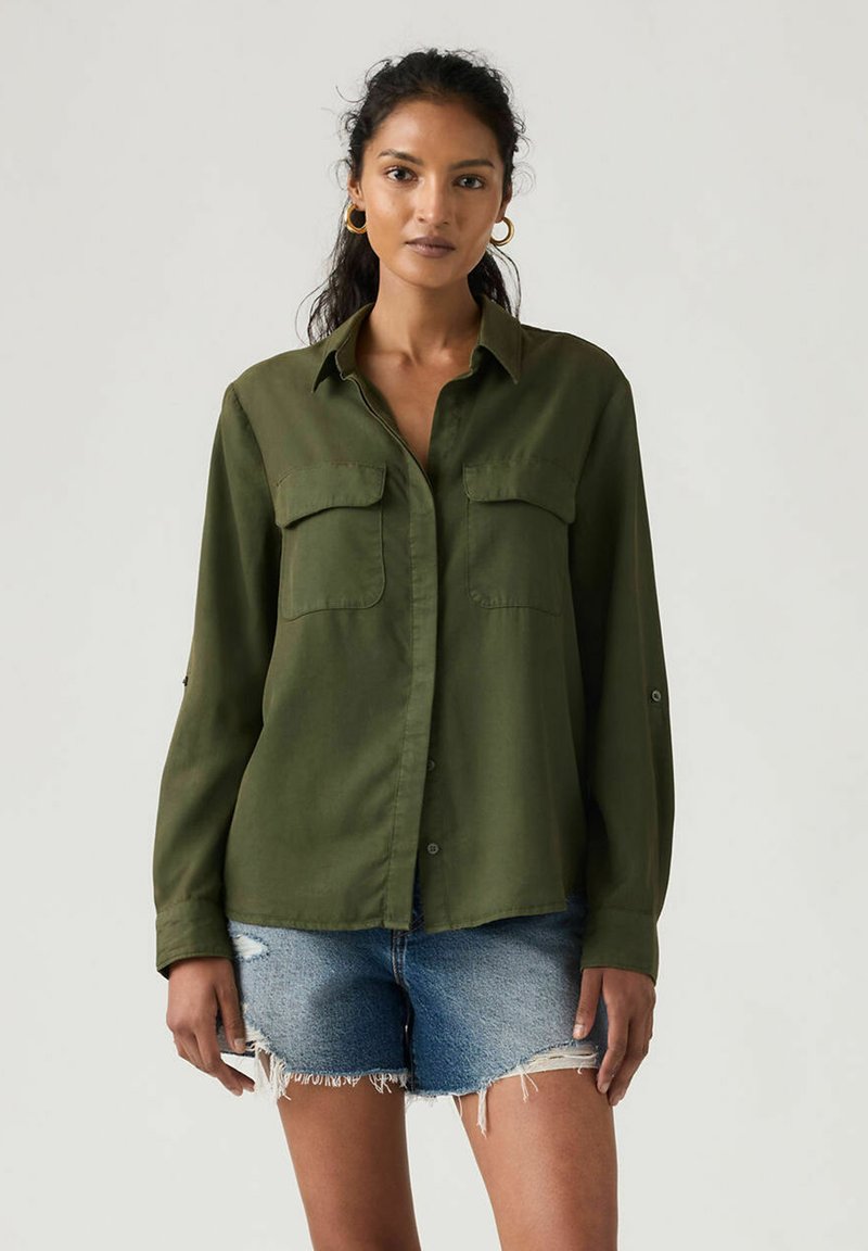 Olivengrøn skjorte med knapper, to brystlommer, let stof, lange ærmer og op rullede manchetter. Bruges med slidte denimshorts.