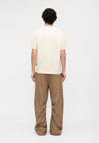 Άτομο με σγουρά σκοτεινά μαλλιά, φορώντας ένα απλό εκρού κοντομάνικο πουκάμισο και χαλαρά καφέ παντελόνια, στέκεται με την πλάτη στο λευκό φόντο.