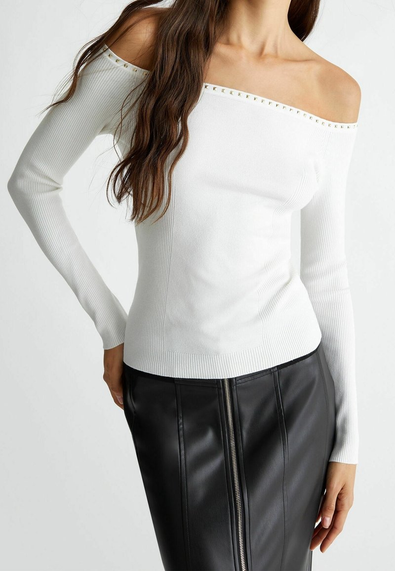 Haut blanc côtelé à épaules dénudées avec des accents de clous dorés au niveau du col, associé à une jupe en cuir noire ajustée avec une fermeture éclair frontale.
