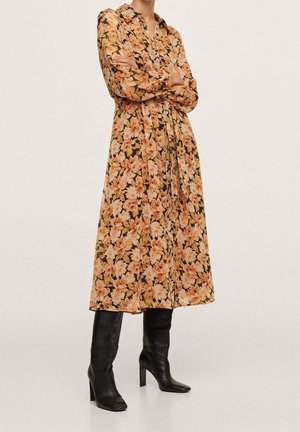 Personne portant une robe fleurie à manches longues boutonnée, dans des tons orange et beige, avec des bottes noires montantes à talons, debout les bras croisés.