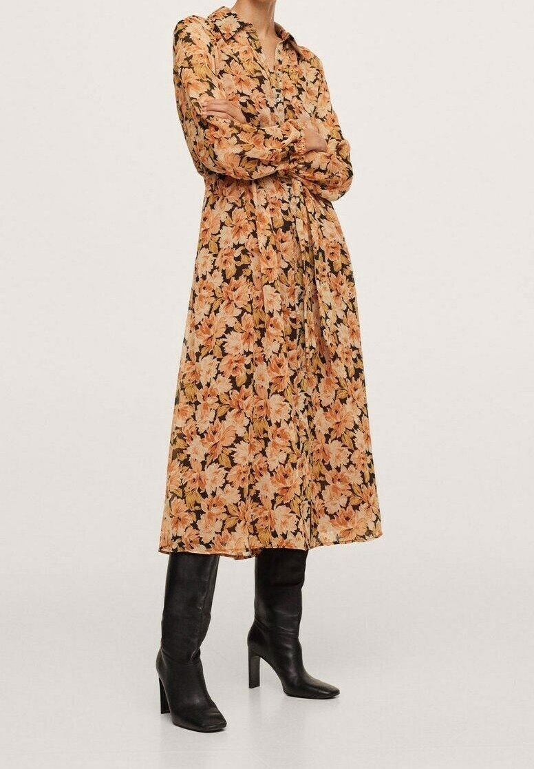 Personne portant une robe fleurie à manches longues boutonnée, dans des tons orange et beige, avec des bottes noires montantes à talons, debout les bras croisés.