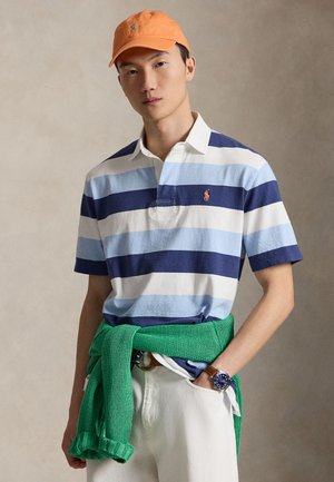 Jeune homme portant une casquette orange, un polo rayé bleu et blanc, un pantalon blanc, un pull vert noué autour de la taille et une montre-bracelet marron.