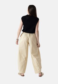Mujer de espaldas vestida con una camiseta negra sin mangas, pantalones beige de pierna ancha con cintura elástica y zapatos beige sin cordones.