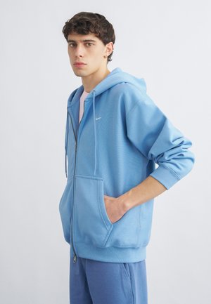 Noor mees lühikeste tumedate lokkis juustega, kandes helehalli lukuga tuultõrjejakki ja sobivaid dressipükse, üks käsi taskus.