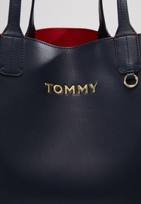 Granatowa skórzana torba tote o gładkiej fakturze, z czerwoną podszewką i złotym logo "TOMMY" z przodu. Ustrukturyzowany kształt z podwójnymi uchwytami.