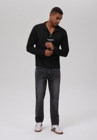 Svart quarter-zip genser med teksturert stoff og hvit logo; kombinert med mørkegrå jeans og hvite sneakers med blå detaljer.