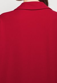 Blouse rouge avec un col cranté et une texture lisse, présentant un design uni sans motifs ni éléments métalliques visibles.