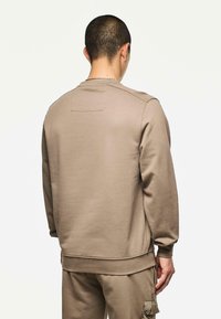 Uomo con capelli corti scuri che indossa un felpa beige e pantaloni cargo abbinati, in piedi con la schiena rivolta contro uno sfondo chiaro e semplice.
