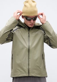 Άνδρας που προσαρμόζει μπεζ πλεκτό καπέλο, φοράει χακί πράσινο μπουφάν Columbia με τσέπες με φερμουάρ και σκούρα γυαλιά ηλίου, μπροστά σε απλό φόντο.