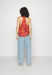 Top halter vermelho com padrão de paisley e costas abertas, combinado com jeans de perna larga azul claro e sapatos de cor creme.