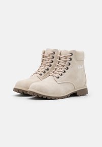 Botines de ante beige con suela de goma marrón, puntera redonda, ojales negros y cordones rosa claro. Presenta el logo de Fila en el costado.