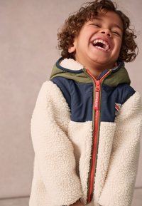 Fleecová bunda s krémovým telom, námorníckymi ramennými panelmi, zelenou kapucňou a jasne oranžovým zipsom. Textúrovaná tkanina s hravou potlačou.