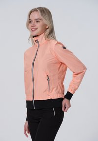 Kevyt, persikanvärinen takki, jossa on korkea kaulus, vetoketjullinen etuosa, sivutaskut ja elastiset hihansuut. Saattaa olla sileä tekstuuri ja moderni muotoilu.