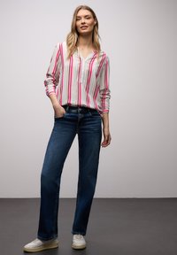 Femme debout, une main dans la poche, portant un chemisier à manches longues rayé rose, blanc et bleu, un jean bleu et des baskets blanches.