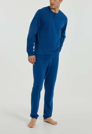 Uomo in piedi a piedi nudi che indossa una maglia blu a maniche lunghe con sottili righe e pantaloni blu coordinati, davanti a uno sfondo semplice.