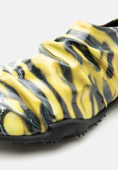 Κοντινή λήψη ενός παπουτσιού με κίτρινο και μαύρο κυματιστό μοτίβο, με υφή στην σόλα και ανυψωμένες ράβδους στην ανώτερη επιφάνεια.