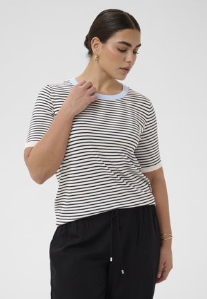 Donna con capelli scuri raccolti in uno chignon, indossa una maglietta a maniche corte a righe bianche e nere e pantaloni neri, si tocca il colletto, guardando verso il basso di lato.