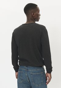 Srajca dolgih rokavov temno sive barve z okroglim izrezom, v kombinaciji z modrimi jeans hlačami. Material se zdi mehak, kroj pa je sproščen.