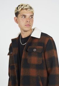 Geruite wollen shirtjas met een bruin en zwart patroon, witte nepbont kraag, twee voorzakken en knoopsluitingen.