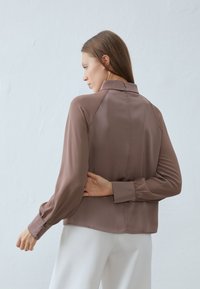 Brown, long-sleeve blouse with a structured collar and gathered cuffs. Smooth fabric with a sleek back design and a button closure.
