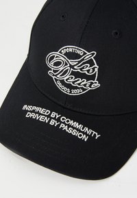 Black baseball cap with embroidered white logo reading "Les Deux Sporting Goods 2024" and text "Inspired by community driven by passion."