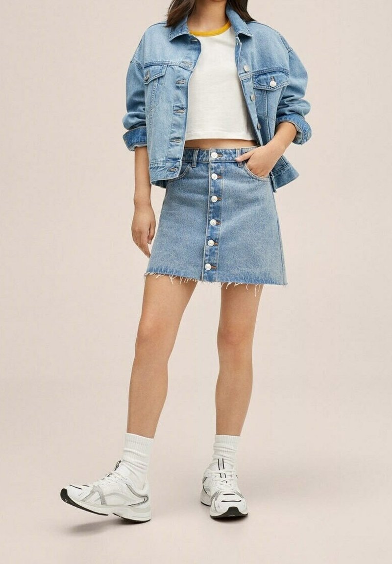 Femme portant une veste en jean claire, un t-shirt court blanc à bords contrastants, une jupe en jean boutonnée devant, des chaussettes blanches et des baskets blanches.