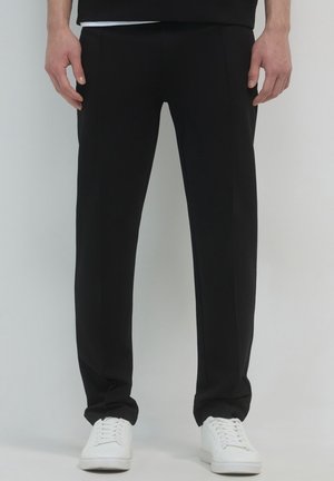 Schwarze, maßgeschneiderte Hose aus einem glatten Stoff, mit geradem Schnitt, subtilen Falten und seitlichen Taschen, kombiniert mit weißen Sneakern.