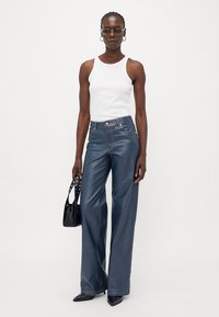 Hoge taille, wijde spijkerbroeken van donkerblauwe denim, gecombineerd met een witte tanktop; model houdt een zwarte handtas met accentstuds vast.