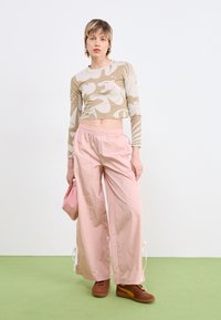 Beige krūšturis ar baltu ziedu rakstu, kombinēts ar brīviem rozā biksēm un brūnām kedām. Modelis tur mazu rozā soma.