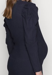 Mörkblå klänning med texturerad tyg, långa ärmar och puffdetaljer på axlarna. Åtsittande siluett framhäver midjan.