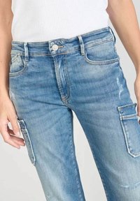 Vista frontal de unos pantalones cargo de mezclilla azul claro con bolsillos laterales, llevados por una persona que viste una camiseta blanca con textura de canalé, cortada a la altura de la cintura.