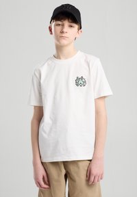 T-shirt bianca in cotone con maniche corte, caratterizzata da un design grafico colorato sul lato sinistro del petto. Abbinata a pantaloni khaki e un berretto nero.