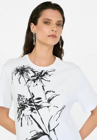Tricou alb cu un design floral de culoare neagră. Supravestiment cu mâneci scurte și decolteu rotund; textură fină de bumbac.