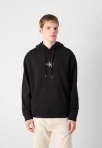 Sudadera con capucha negra fabricada en mezcla de algodón, con un bolsillo frontal y un logo blanco. El modelo lleva pantalones de color claro y está de pie frente a un fondo gris.
