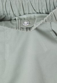 Light green fabric with a smooth texture, featuring an elastic waistband and a button closure. Close-up view highlighting stitching details.