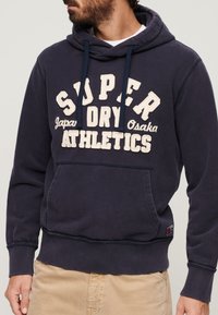 Man draagt een donkerblauwe hoodie met de tekst "Super Dry Athletics" en "Japan Osaka", gecombineerd met beige broek, kijkt zijwaarts.