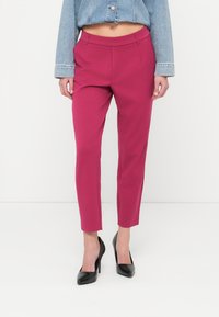 Παντελόνια σε φούξια από ελαστικό ύφασμα με ελαφρώς ελαστικό υλικό, διαθέτοντας λείο ζωνάρι μέσης και εμπρόσθιες τσέπες, σε συνδυασμό με μαύρες ψηλοτάκουνες γόβες.