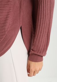Dzianinowy sweter w kolorze przybrudzonego różu, z pionowymi i poziomymi fakturami rib, długimi rękawami oraz bocznymi rozcięciami dla większej swobody ruchów.