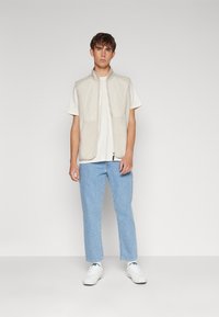 Beige ermeløs vest med glidelås foran og to lommer, sammen med en hvit T-skjorte og lyseblå jeans med rette ben, iført hvite sneakers.