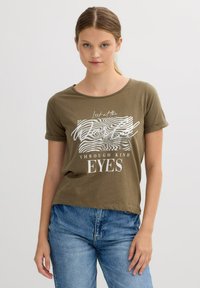 Camiseta verde oliva de mangas cortas enrolladas y escote redondo. Presenta texto gráfico en blanco y líneas onduladas. Material de algodón suave.