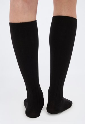 Jambes portant des chaussettes noires côtelées montant jusqu'aux genoux, vues de dos sur un fond clair uni.