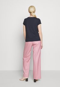 Nainen, jolla on vaaleat poninhännät, pukeutuneena mustaan lyhythihaiseen toppiin, vaaleanpunaisiin leveisiin housuihin ja beigekalvoisiin korkokenkiin, seisoo yksinkertaista taustaa vasten.