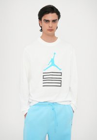 Balta garo piedurkņu krekls ar tirkīza Jumpman logo un melnām horizontālām līnijām. Apakšā valkā gaiši zilos sporta bikses.