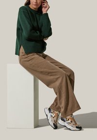 Maglione verde a maglia, pantaloni larghi marroni e sneakers con motivo e accenti bianchi e neri, poggiati su un pedestal bianco.