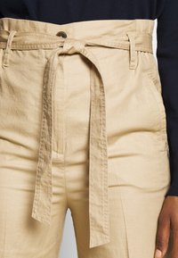 Beige Hochwaist-Hosen mit einem Stoffgürtel, ausgestattet mit Seitentaschen und einer glatten Textur. Mit einem vorderen Knopfverschluss akzentuiert.
